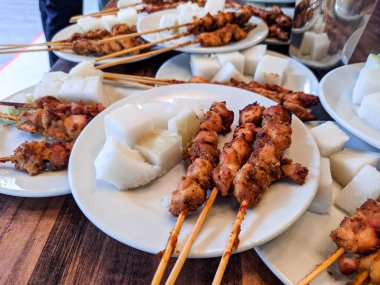 Beyaz bir tabakta servis edilen ve ahşap bir masada düzgünce hazırlanmış pirinç keki ile satay tavuğu.