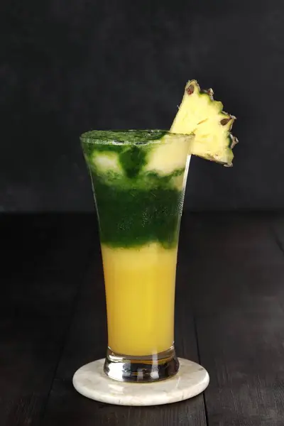 pineapple juice with vegetables and pineapple chunks garnish. has yellow and green gradations. served in a clear glass with a white and dark background