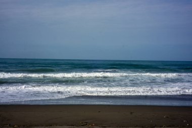 Güçlü köpüklü deniz dalgaları bulutlu mavi gökyüzüne doğru yuvarlanıyor ve sıçrıyor.