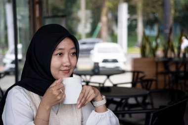 Endonezyalı kadın tesettürcü kafede oturuyor. İş kadını kahve dükkanında çalışıyor ve elinde bir fincan kahve tutuyor. Siyah başörtüsü ve beyaz pıhtı giyiyor.