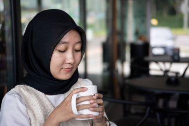 Endonezyalı kadın tesettürcü kafede oturuyor. İş kadını kahve dükkanında çalışıyor ve elinde bir fincan kahve tutuyor. Siyah başörtüsü ve beyaz pıhtı giyiyor.