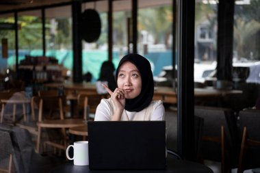 Kafede otururken dizüstü bilgisayar kullanan mutlu bir kadın görüntüsü. Kafede oturan ve dizüstü bilgisayarla çalışan tesettürlü genç Asyalı kadın..