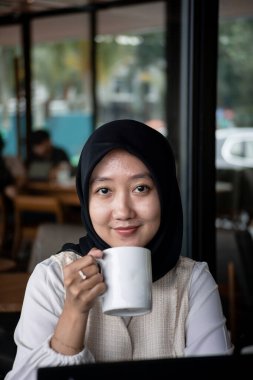 Güzel genç bir kadın bir dizüstü bilgisayarda çalışıyor ve gülümsüyor ve bir kafede oturmuş kahve içerken düşünüyor..