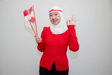 Beyaz tesettürlü, kırmızı kazaklı bir kadın. Endonezya 'nın bağımsızlık gününü kutluyoruz. Elinde bayrak ve el işareti var. Kameraya bakıyorum. Beyaz arkaplanda izolasyon.