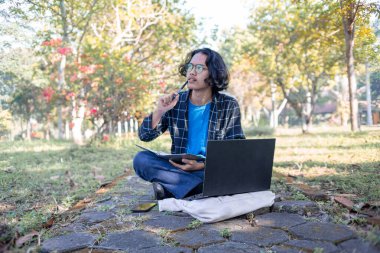 Bir erkek öğrenci kampüs bahçesinde dizüstü bilgisayarla oturuyor. Düşünceli bir ifadeyle.