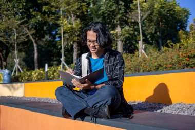 Bir erkek öğrenci parkta oturuyor ve bir kitap yazıyor..