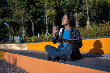 Erkek öğrenci parkta oturuyor ve elinde düşünceli bir ifadeyle bir kitap tutuyor..