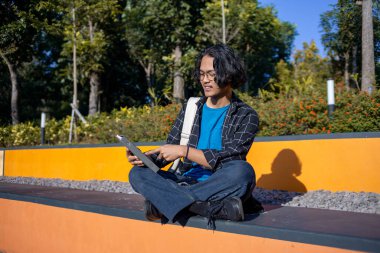 Bir erkek öğrenci parkta oturuyor ve bir kitap okuyor..