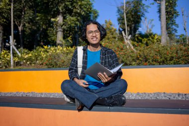 Bir erkek öğrenci parkta oturuyor ve gülümserken bir kitap tutuyor..