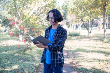 Parkta gömlek ve gözlük takan bir erkek öğrenci bir kitap tutuyor..