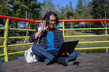 Gömlek ve gözlük takan bir erkek öğrenci parkta oturuyor ve merhaba diyor..