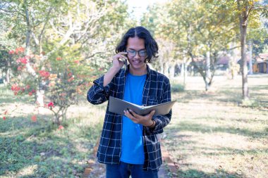Ekose gömlek ve gözlük takan bir erkek öğrenci parkta duruyor. Telefona cevap veriyor ve elinde kitap tutuyor..