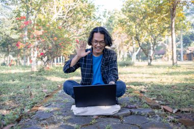 Ekose gömlek ve gözlük takan bir erkek öğrenci parkta dizüstü bilgisayarıyla oturuyor. Bilgisayara merhaba de.