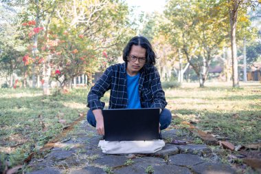 Ekose gömlek ve gözlük takan bir erkek öğrenci parkta dizüstü bilgisayarıyla oturuyor. Düşünceli bir ifadeyle..