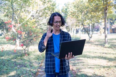 Ekose gömlek ve gözlük takan bir erkek öğrenci parkta duruyor. Bilgisayara merhaba de.