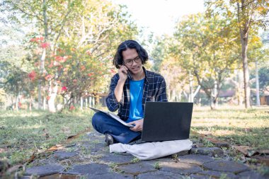 Ekose gömlek ve gözlük takan bir erkek öğrenci parkta oturmuş laptopunun önündeki telefona cevap veriyor..