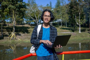 Ekose gömlek ve gözlük takan bir erkek öğrenci parkta gülümseyen bir ifadeyle duruyor..