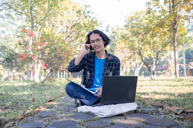 Ekose gömlek ve gözlük takan bir erkek öğrenci parkta oturmuş laptopunun önündeki telefona cevap veriyor..