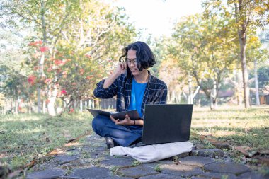 Ekose gömlek ve gözlük takan bir erkek öğrenci parkta oturmuş laptopunun önündeki telefona cevap veriyor..