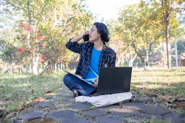 Ekose gömlek ve gözlük takan bir erkek öğrenci parkta oturmuş laptopunun önündeki telefona cevap veriyor..