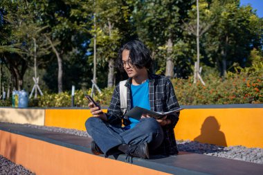 Ekose gömlek ve gözlük takan bir erkek öğrenci parkta oturmuş gülümsüyor ve elinde kitap tutuyor..