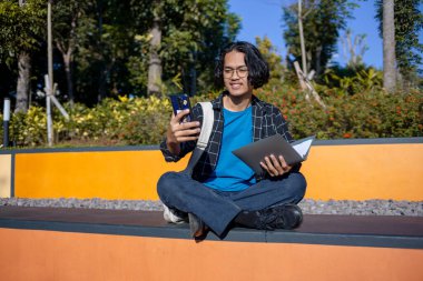 İşaretli gömlek ve gözlük takan bir erkek öğrenci parkta oturup telefonuyla selfie çekiyor..