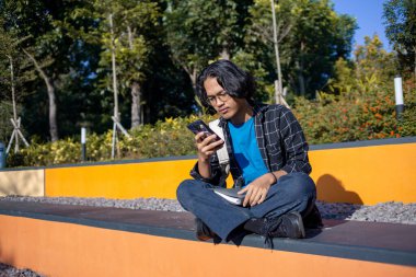 Ekose gömlek giyen bir adam, cep telefonuyla çeşitli ifadelerle oynarken parkta oturur..