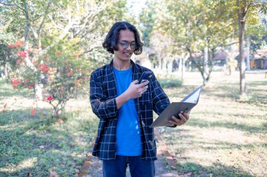 Ekose gömlek ve gözlük takan bir erkek öğrenci parkta durmuş görev kitabının fotoğraflarını çekiyor..