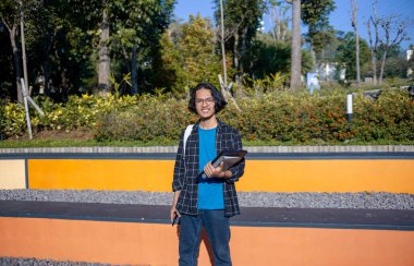 Ekose gömlek ve gözlük takan bir erkek öğrenci parkta elinde bir kitapla durup gülümsüyor..