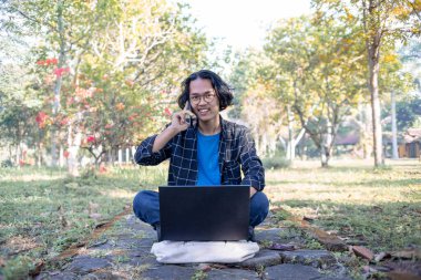 Ekose gömlek giyen bir erkek öğrenci parkta oturmuş mutlu bir ifadeyle bir telefon alıyor..