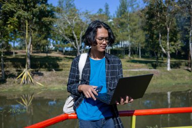 Ekose gömlek giyen bir erkek öğrenci elinde dizüstü bilgisayarla parkta duruyor..