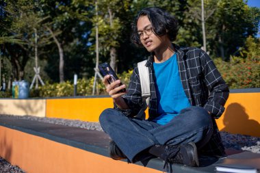 Ekose gömlek giyen bir adam, cep telefonuyla çeşitli ifadelerle oynarken parkta oturur..