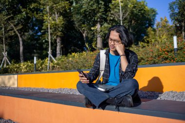 Ekose gömlek giyen bir adam, cep telefonuyla çeşitli ifadelerle oynarken parkta oturur..