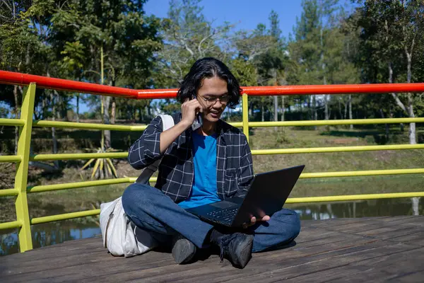 Ekose gömlek giyen bir erkek öğrenci parkta oturmuş mutlu bir ifadeyle bir telefon alıyor..