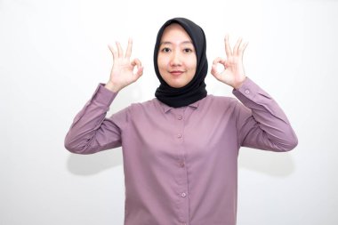 Endonezyalı, tişörtlü ve tesettürlü bir Asyalı kadının portresi. Bir eli gözünün üzerinde kameraya parmaklarından bakıyor. Anlaşma işareti veriyor. Beyaz arkaplanda izole edilmiş