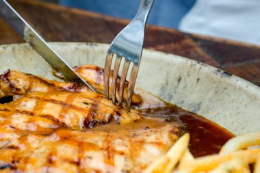 Bir kadın bir restoranda nefis bir tavuk bifteği ve patates kızartmasının tadını çıkarıyor, nefis bir deseni olan krema renkli bir tabakta servis ediliyor. Yemek şık bir ahşap masaya konur.