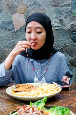 Siyah tesettürlü ve mavi gömlekli bir kadın taştan bir duvarın önünde yemeğin tadını çıkarıyor. Rahatlamış ve yemeğine odaklanmış görünüyor. Dokulu, doğal bir zemin üzerine kurulmuş.