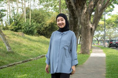 Siyah tesettürlü ve mavi gömlekli neşeli bir kadın güzel bir park kaldırımının kenarında duruyor, gülümsüyor ve anın tadını çıkarıyor. Dışarıdaki hareketli ortam onun neşeli ifadesini artırıyor.