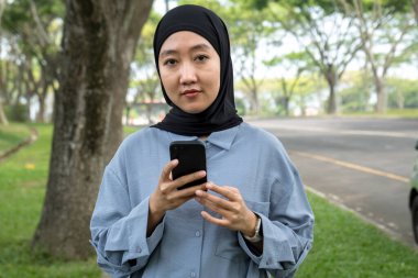 Siyah tesettürlü ve mavi gömlekli neşeli bir kadın güzel bir park kaldırımının kenarında duran akıllı bir telefonu tutarken parlak bir şekilde gülümsüyor. Neşeli ifadesi açık hava manzarası ile tamamlanıyor.