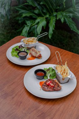 Biftek ve biftek, patates kızartması, taze salata ve ahşap masa üzerindeki beyaz tabakta soslar..