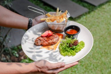 Elleri taze salata, tuzlu sos ve patates kızartması ile sulu biftek tutuyor, beyaz tabakta servis ediliyor, zengin ve lezzetli bir yemek..