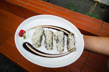 Kızarmış muz tatlısı, üstüne peynir ve çikolata serpiştirilmiş çikolata sosu ve açık havada beyaz bir tabakta çilek..