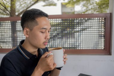 İki eliyle kahve içen genç adam, rahat bir ortamda sıcak bir içecek tutarken rahatlamanın tadını çıkarıyor..