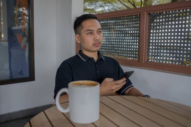 Adam telefonu tutuyor ve elinde bir bardak kahveyle masadan uzaklaşıyor, düşüncelere dalma ve rahatlama anının tadını çıkarıyor..