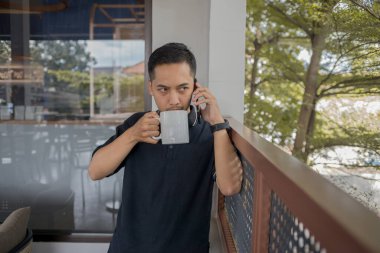 Adam telefonda konuşurken ayakta durup kahve içiyor ve sakin bir ortamda bir anlık rahatlığın tadını çıkarıyor..