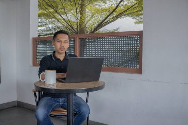 Laptopta çalışan bir adam masanın üzerinde bir bardak kahveyle, sıcak bir kafe atmosferine bakıyor..