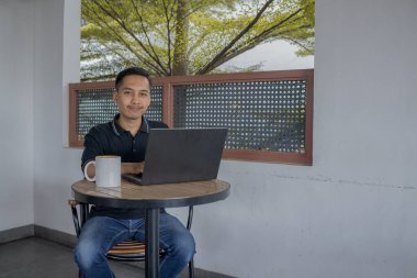 Laptopta çalışan bir adam, masada bir bardak kahveyle, rahat bir kafede beklerken gülümsüyor..