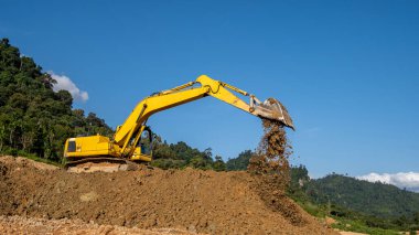 Kazı makinesi, arka planda tropikal ormanlar ve açık mavi gökyüzü olan tepelerin üzerinde toprak kazıp döküyor.