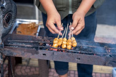 Street vendor grilling skewers of meatball-like food over charcoal clipart