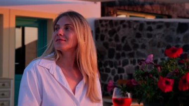 Sarışın kadın akşamları dışarıda dinlenir, beyaz gömlek giyer, şarap kadehi tutar, huzurlu bir anın tadını çıkarır.